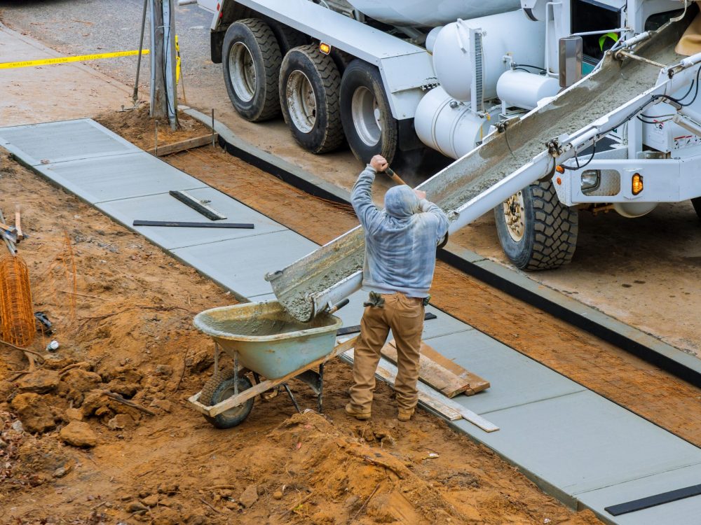 Construction worker pour cement for sidewalk in concrete works with mixer truck with wheelbarrow at construction site filling formwork with cement and gravel
